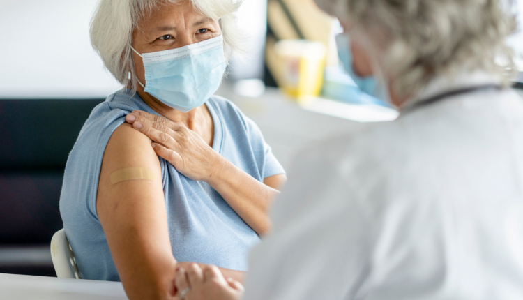 Een mevrouw heeft een pleister op haar arm na een vaccinatie. Ze draagt een mondkapje en heeft grijs haar.